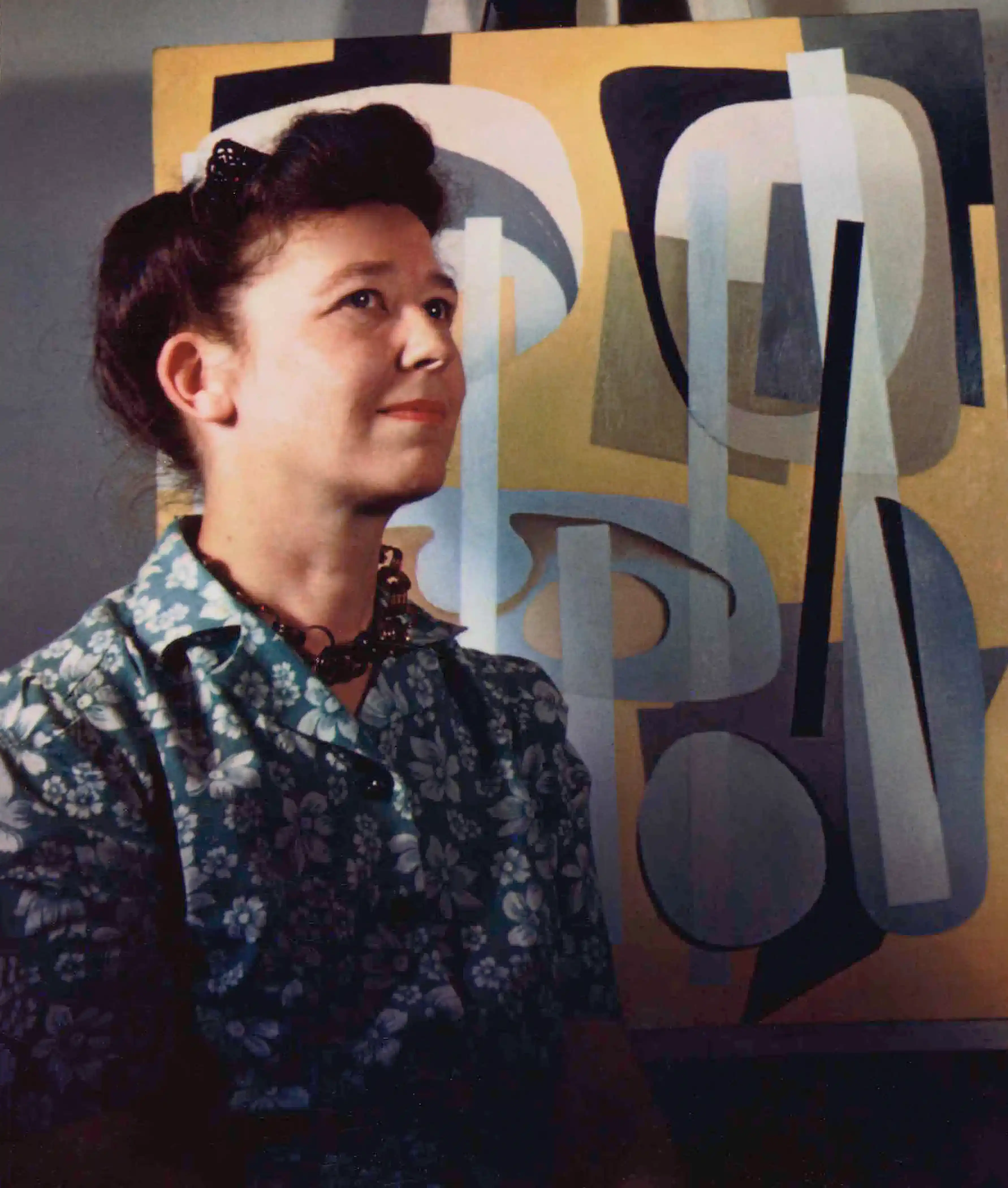 Color portrait photograph from 1946 of a woman with a light skin tone, standing in front of a yellow abstract painting resting on an easel.